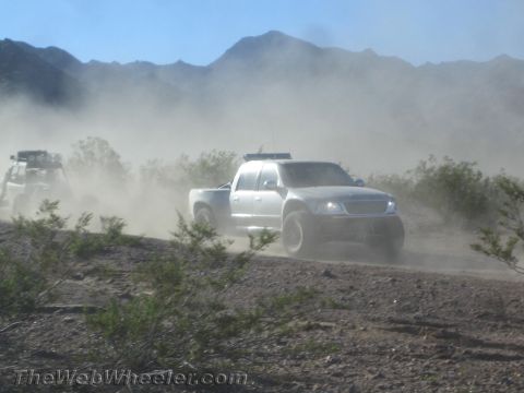 1990 Ford Bronco - white off-road race truck and a buggy speed through a cloud of desert dust with mountains behind.