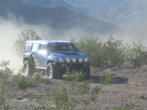 1990 Ford Bronco - blue Hummer H3 with auxiliary lights climbs a dusty desert hillside surrounded by scrub brush and mountains.