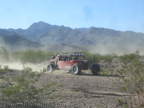 1990 Ford Bronco - red and silver tube-chassis off-road buggy races across open desert flats trailing dust below a mountain range.
