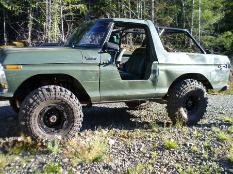 1978 Ford Bronco - Driver-side profile of a lifted flat-green full-size Ford Bronco with the top and doors removed, showing a black roll cage, black bucket se…