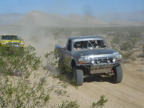 1990 Ford Bronco - gray Ford prerunner race truck numbered 1501 climbs a sandy desert trail, with a yellow race truck following close behind.