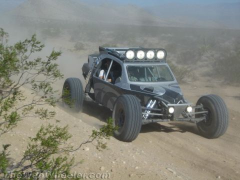 1990 Ford Bronco - black single-seat tube-frame off-road race buggy fitted with four roof-mounted driving lights speeds along a dirt desert road.