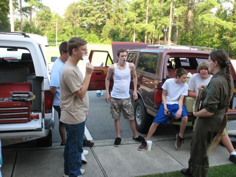 1995 Ford C-150 (Centurion Conversions) - Several young people stand and sit around two SUVs parked in a driveway.