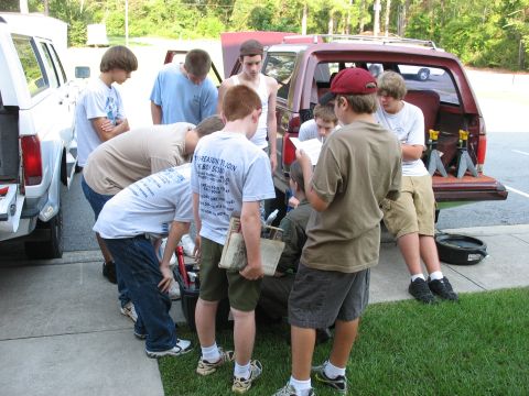 1995 Ford C-150 (Centurion Conversions) - A group of boys are gathered around the open rear of a red SUV, with a white truck visible to the left.