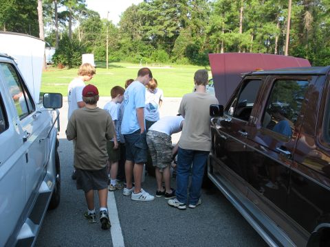 1995 Ford C-150 (Centurion Conversions) - A group of boys inspects the engines of two vehicles with their hoods open on a sunny day.