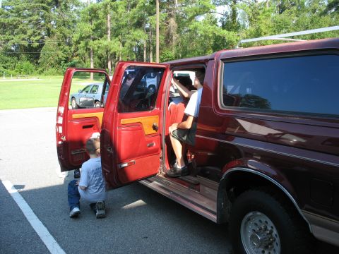 1995 Ford C-150 (Centurion Conversions) - A red SUV has its doors open as children prepare to enter, with another vehicle visible in the background.