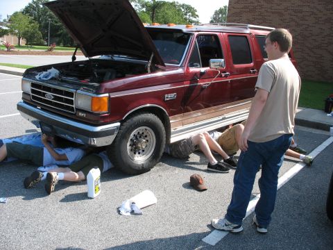 1995 Ford C-150 (Centurion Conversions) - Several individuals are gathered around a maroon Ford F-Series based SUV, with some working underneath it and the hood open.