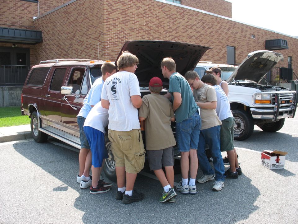 Group of teens huddled around the open hood of a maroon full-size SUV in a parking lot