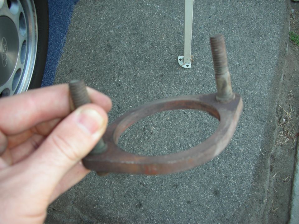 Rusty exhaust flange with two bolts held by a hand, with a car wheel and blue carpet visible