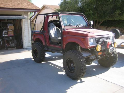 1987 Suzuki Samurai - This red 1987 Suzuki Samurai is equipped with an external roll cage and oversized tires, suggesting off-road modifications.