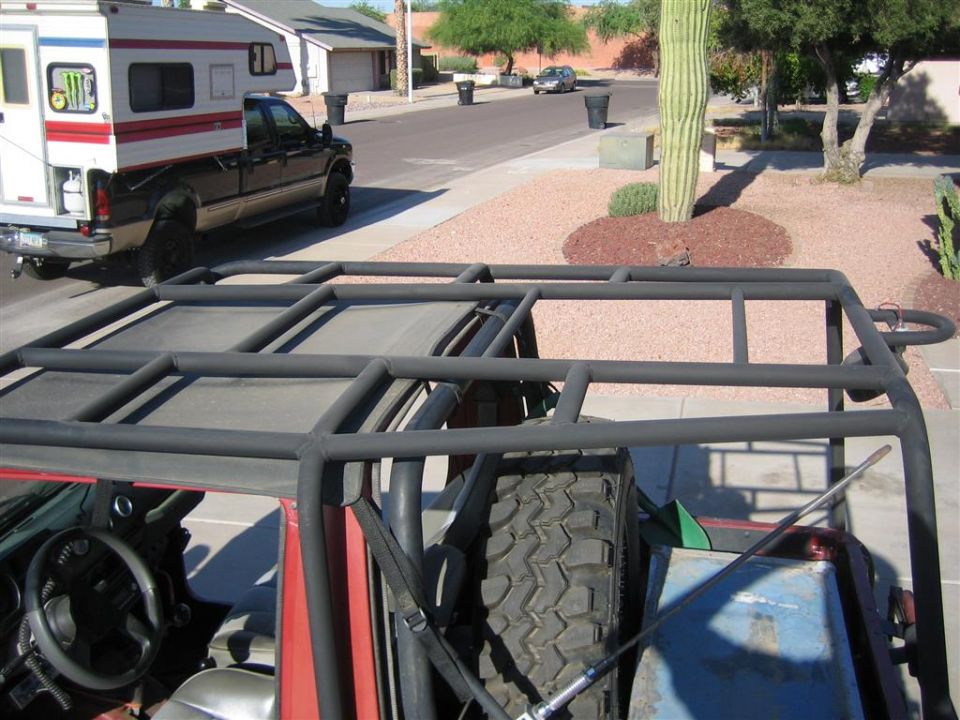 Custom gray tubular roof rack over small SUV with spare tire, soft top, desert street behind
