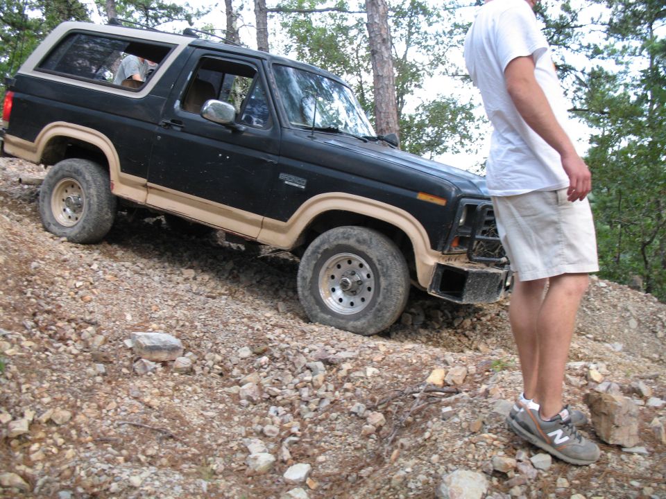 Black and tan two-tone Ford Bronco descending a rocky woodland trail with a man watching