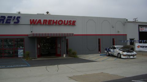 1990 Ford Probe - A race car sits outside a tire warehouse with its hood open, displaying racing decals and the number 65.