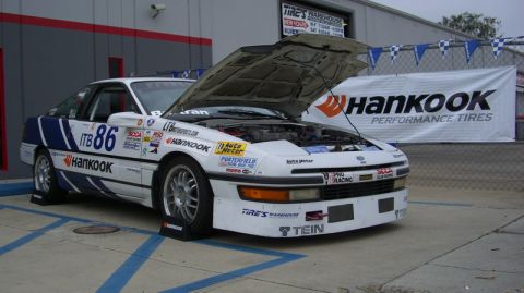 1990 Ford Probe - A white 1990 Ford Probe race car, adorned with sponsor decals and the number 86, is parked with its hood open in front of a building.