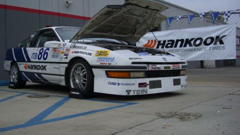 1990 Ford Probe - A white race car with blue stripes and the number 86 is parked with its hood open, displaying various sponsor decals.