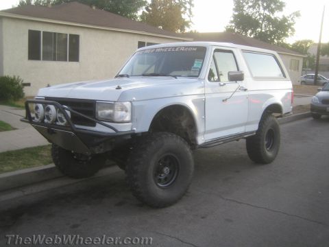 1990 Ford Bronco - white lifted Ford Bronco on large off-road tires with a tubular brush bar and round lights is parked at the curb of a residential street.