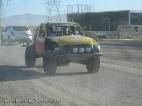 1990 Ford Bronco - yellow and red open-cab off-road truck fitted with round driving lights drives along a gravel staging road past a parked car and a trailer.