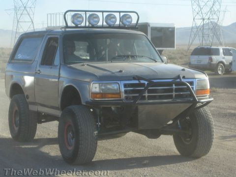 1990 Ford Bronco - gray lifted Ford Bronco prerunner with a four-light roof bar and tubular front bumper sits in a desert staging area near power-line towers.