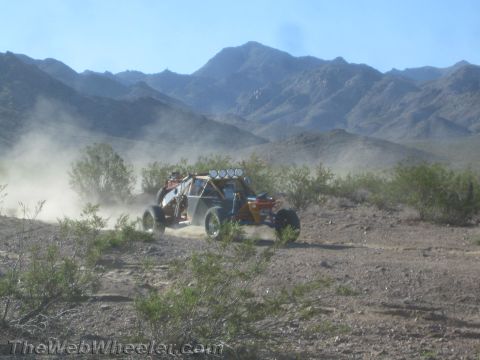 1990 Ford Bronco - orange and blue tube-chassis off-road race buggy speeds along a desert trail, raising dust against a backdrop of rugged mountains.