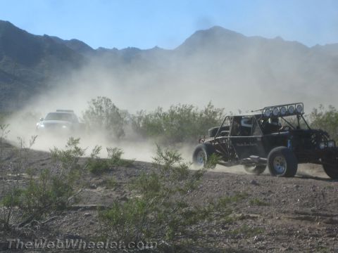 1990 Ford Bronco - tube-frame off-road race buggy with a roof light bar races down a dusty desert trail as a white chase vehicle approaches from the opposite…