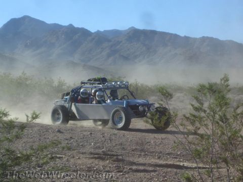 1990 Ford Bronco - two-seat tube-frame off-road race buggy with a roof light bar speeds along a dusty desert trail, mountains rising in the hazy background.