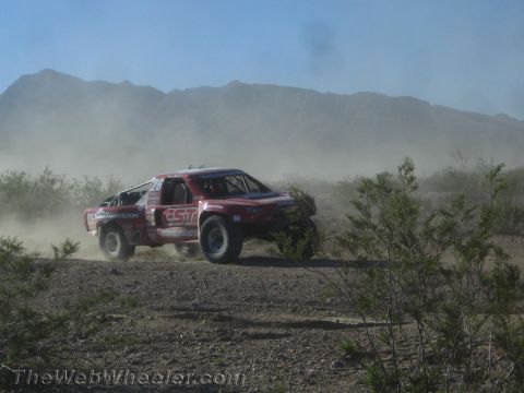 1990 Ford Bronco - red desert race truck speeds along a dusty trail, trailing a cloud of dust with mountains in the distance.