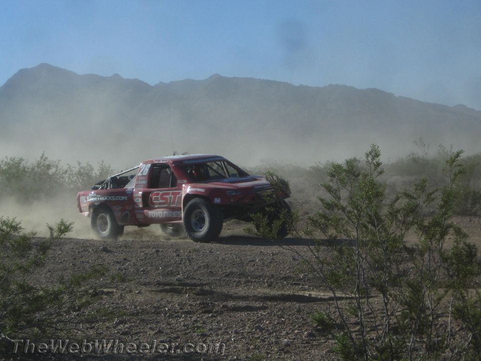 Red off-road race truck kicking up dust on a desert trail with mountains behind