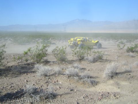 1990 Ford Bronco - yellow off-road buggy crosses a flat desert plain dotted with brush, power lines and distant mountains behind.