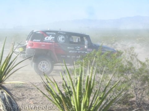 1990 Ford Bronco - red and black Toyota-badged off-road race truck catches air on a dusty desert course.