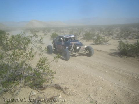 1990 Ford Bronco - open tube-frame off-road race buggy drives along a sandy desert trail lined with brush.