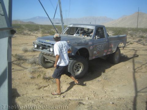 1990 Ford Bronco - man in a white shirt and cap walks past a parked gray Ford race truck marked 1501 beneath desert power lines.