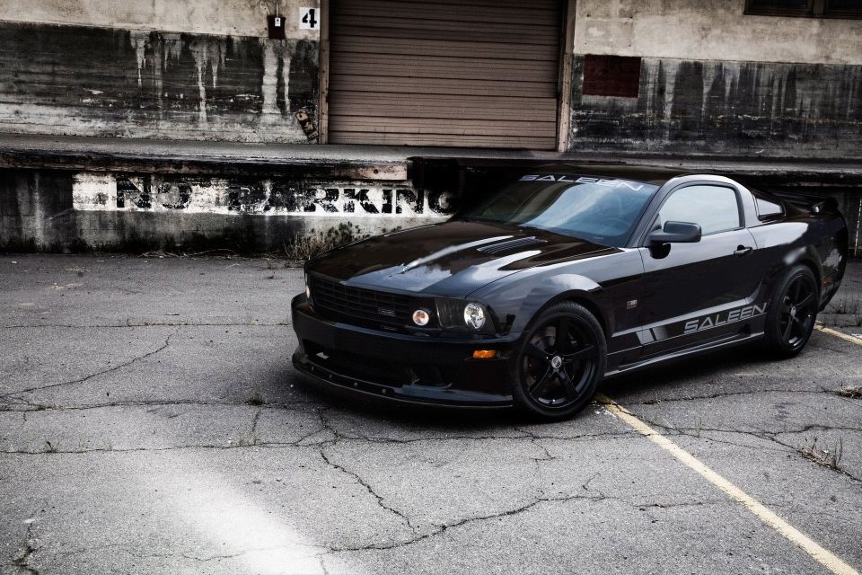 Black Saleen Mustang parked on cracked asphalt in front of a weathered building with a garage door
