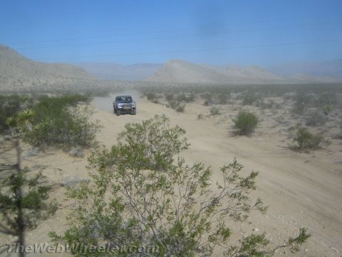 1990 Ford Bronco - dark truck travels a dirt road across open desert with mountains in the distance and brush in the foreground.