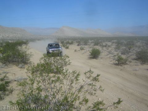 1990 Ford Bronco - dark truck approaches along a sandy desert trail, kicking up dust against a backdrop of hills.