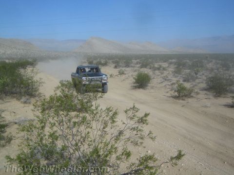 1990 Ford Bronco - blue-gray Ford truck drives a sandy desert road, trailing dust with mountains in the distance.