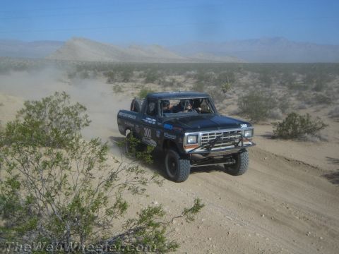 1990 Ford Bronco - dark blue Ford race truck marked 300 drives a desert dirt road, raising dust with mountains behind.