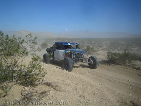 1990 Ford Bronco - blue and silver tube-frame off-road race buggy crests a sandy desert rise trailing dust.