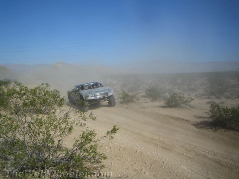 1990 Ford Bronco - silver trophy-style off-road race truck launches over a dusty desert road amid heavy dust.
