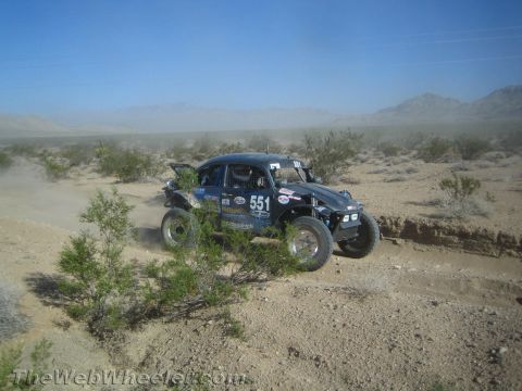 1990 Ford Bronco - black Volkswagen Baja Bug race car marked 551 powers along a rutted desert trail trailing dust.