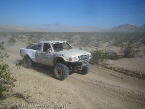 1990 Ford Bronco - white Ford Ranger-style prerunner race truck drives a sandy desert road with mountains in the distance.