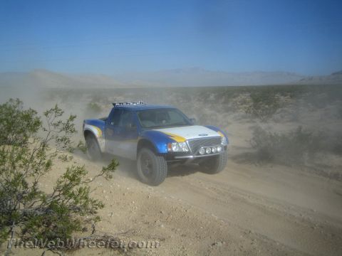 1990 Ford Bronco - blue, white and yellow Ford trophy-style race truck speeds along a dusty desert trail.