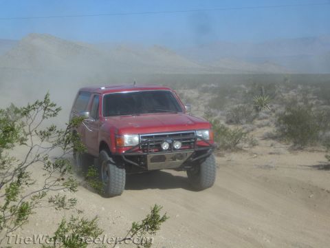 1990 Ford Bronco - red Ford Bronco fitted with a brush guard and lights sits on a sandy desert trail with hills behind.