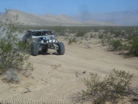 1990 Ford Bronco - silver and blue tube-frame off-road race buggy drives a sandy desert trail with mountains in the distance.