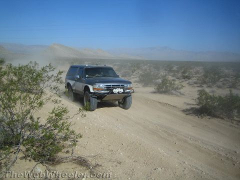 1990 Ford Bronco - gray lifted full-size SUV kicks up dust on a sandy desert trail with mountains in the distance.