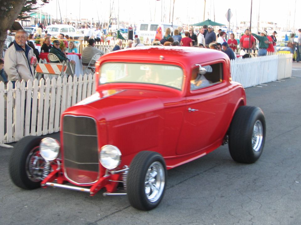 Red hot rod coupe with large rear tires, chrome wheels, and exposed front suspension, viewed from a 3/4 front angle