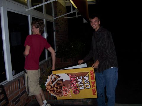 1991 Ford Bronco - Two young men carry a large promotional sign for a "Giant King Cone" ice cream treat outside a building at night.