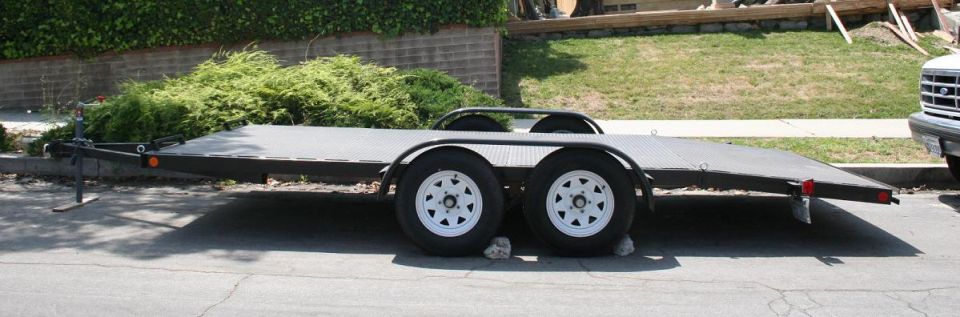 Car trailer with dual axles and white wheels parked on a street, a white truck is partially visible on the right