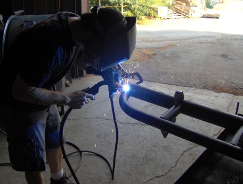 1978 Ford Bronco - A person in a welding mask is actively welding a section of a black metal frame.