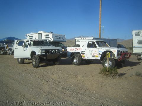 1990 Ford Bronco - Two white off-road race trucks, one numbered 302, sit parked in a dusty desert pit beside RVs.