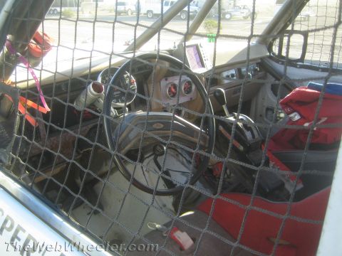 1990 Ford Bronco - stripped interior of a race truck viewed through a window net, showing the steering wheel, gauges and a red seat.